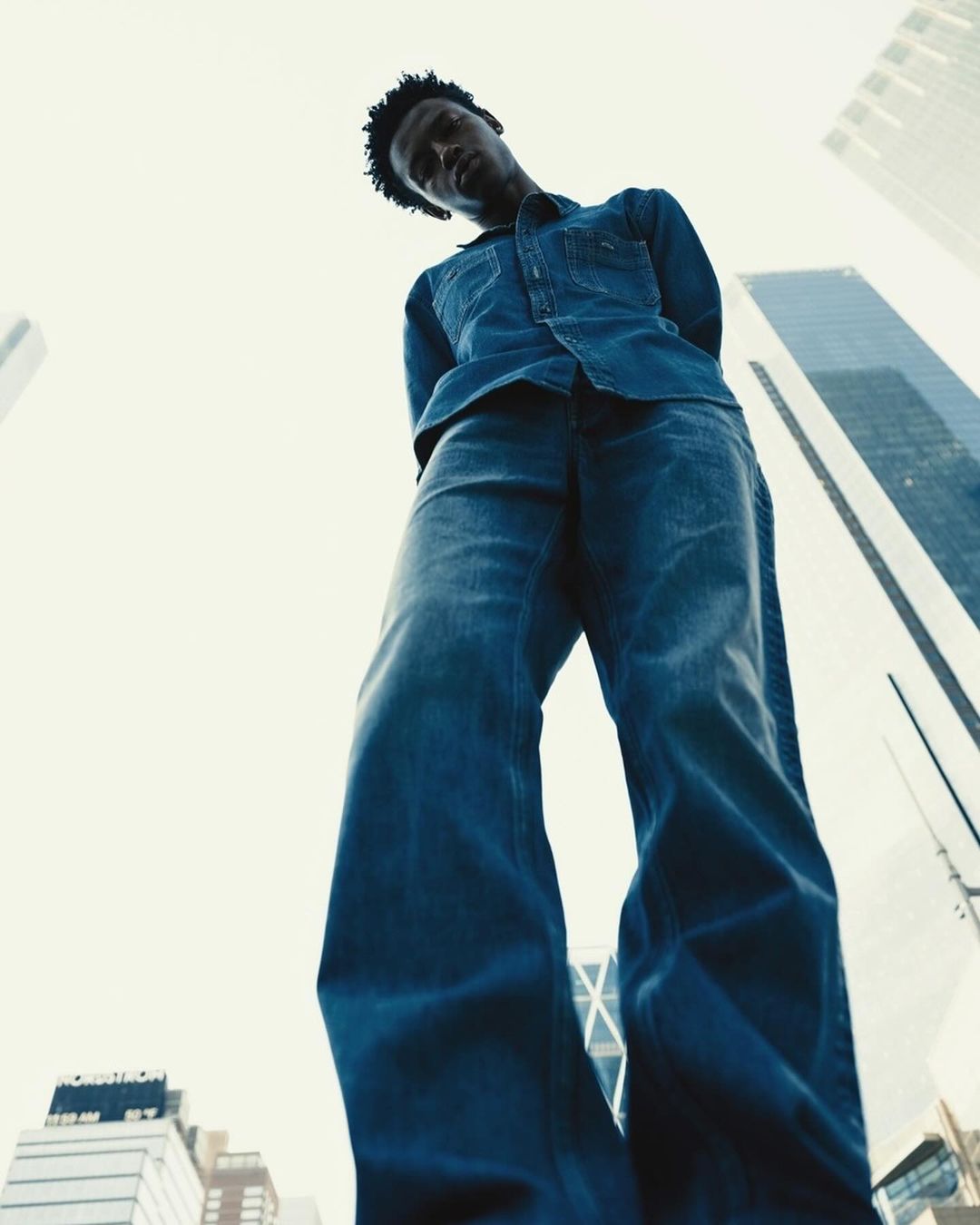 Wide angle shot of an African-American man in a denim shirt and jeans looking down in an urban setting with skyscrapers in the background.