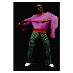 Black woman wearing a pink sweater and patterned green trousers posing against a solid black background.