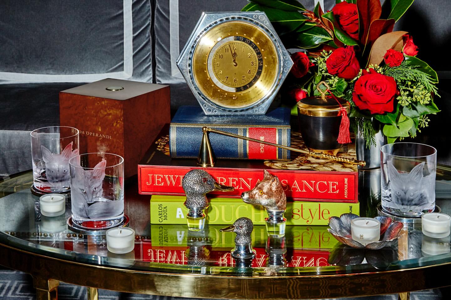 Still life arrangement featuring vintage clock, floral bouquet, books, and decorative animal head bottle stoppers on a glass table.
