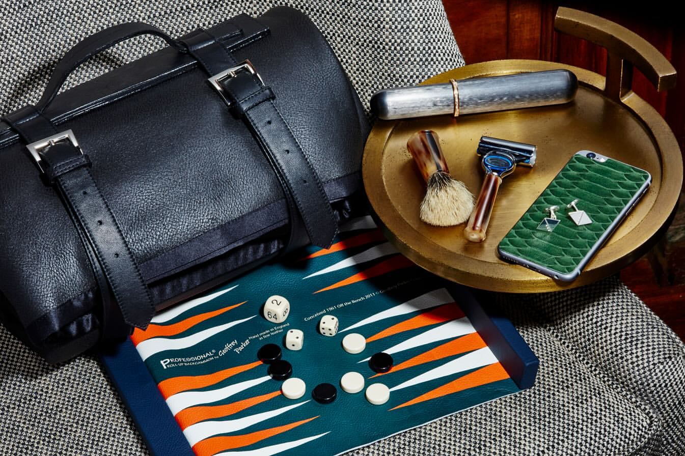 Elevated still life of men's accessories and grooming items: leather bag, backgammon set, razor, shaving brush, cuff links, phone, and travel case.