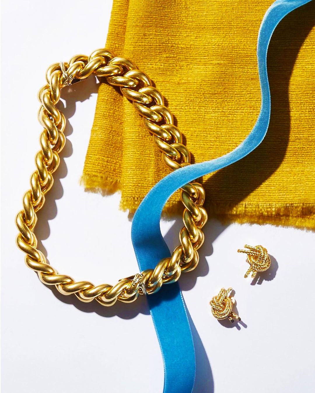 Gold chain necklace and stud earrings are displayed with a curled blue velvet ribbon, and a yellow fabric square on a white background.
