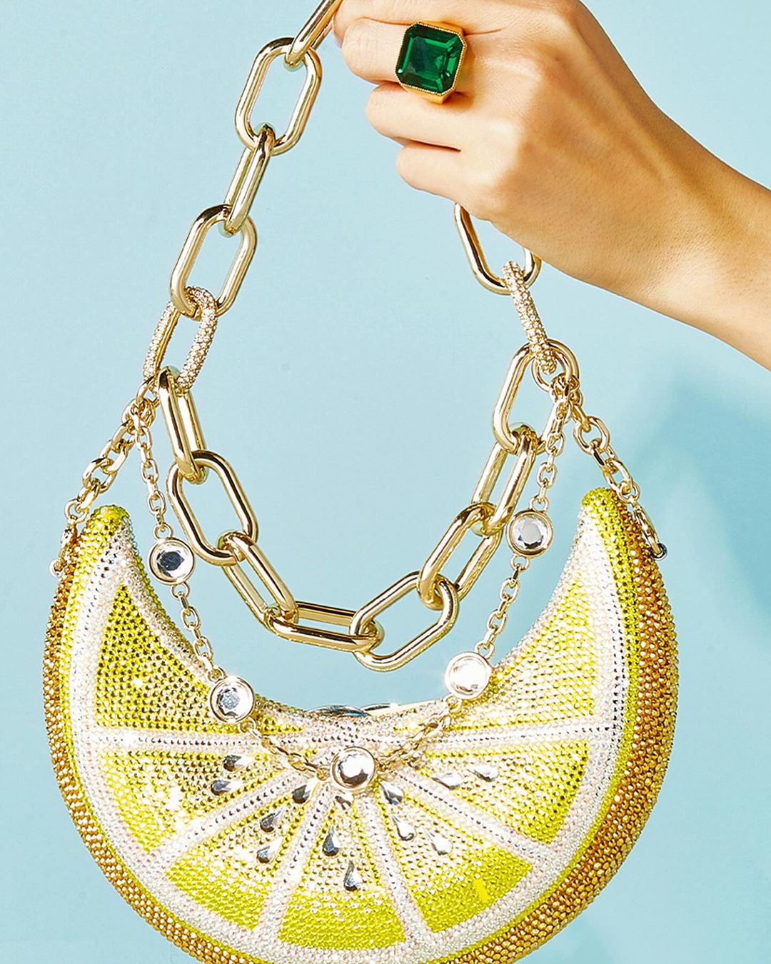 Rhinestone lemon wedge handbag with gold chain straps on a blue background, held by a hand wearing an emerald ring. Fashion accessory.