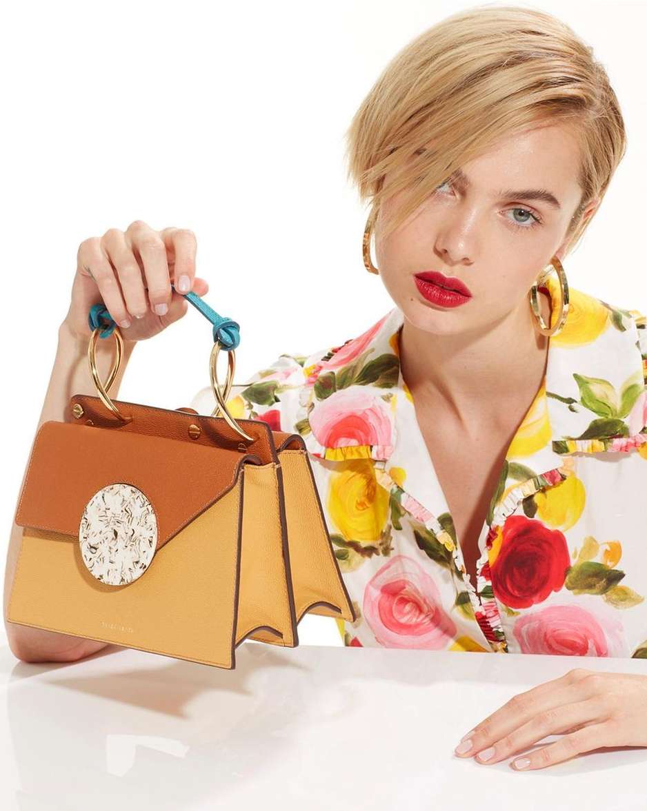 Woman showcases a tan leather handbag with a geometric design against a white backdrop, wearing floral shirt, gold hoop earrings, and red lipstick.