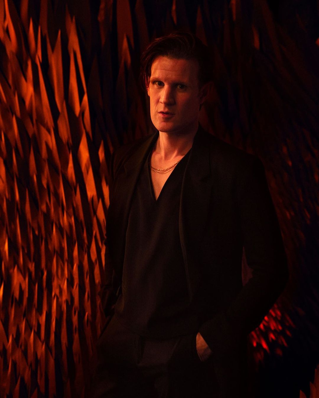Headshot of Matt Smith posing in a black suit jacket against a textured orange wall in a dimly lit indoor environment.
