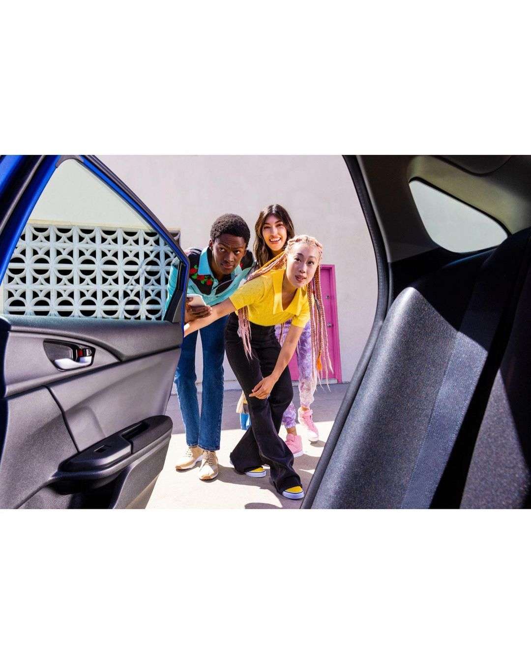 Three young adults peek inside a blue car. Car door is open on a bright, sunny day. Two women and a man are smiling.
