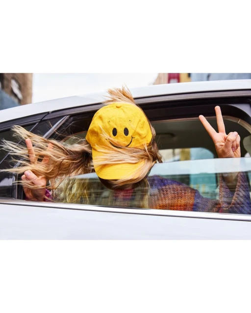 Person in car with a yellow smiley face hat and blonde hair showing peace signs from the car window with urban background.