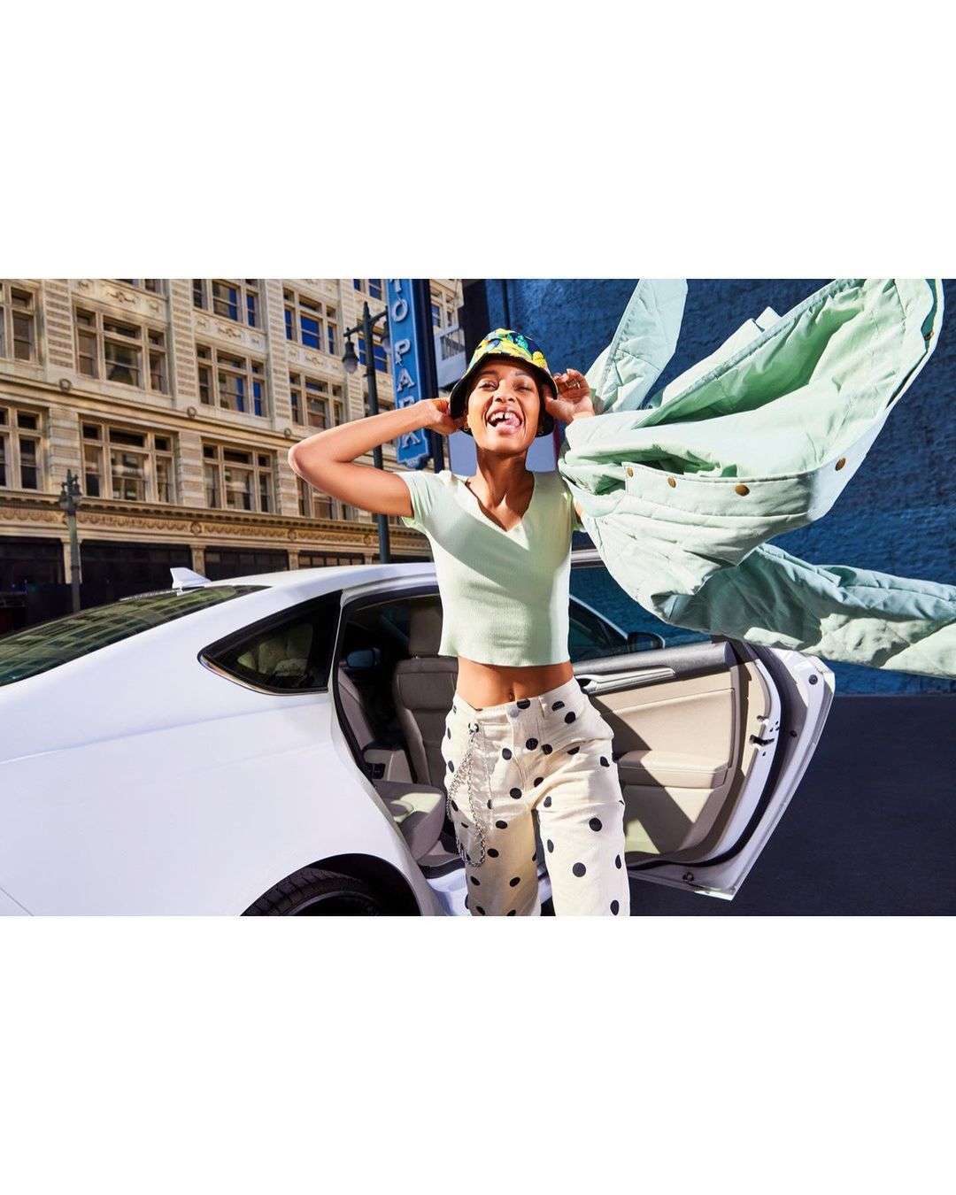 A happy adult woman with a hat exiting a white car on a city street. She's wearing patterned pants and a light green shirt.