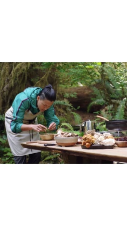 Woman in a forest prepares food at a wooden table with mushrooms, a teapot, and cooking pots, surrounded by ferns and mossy trees.