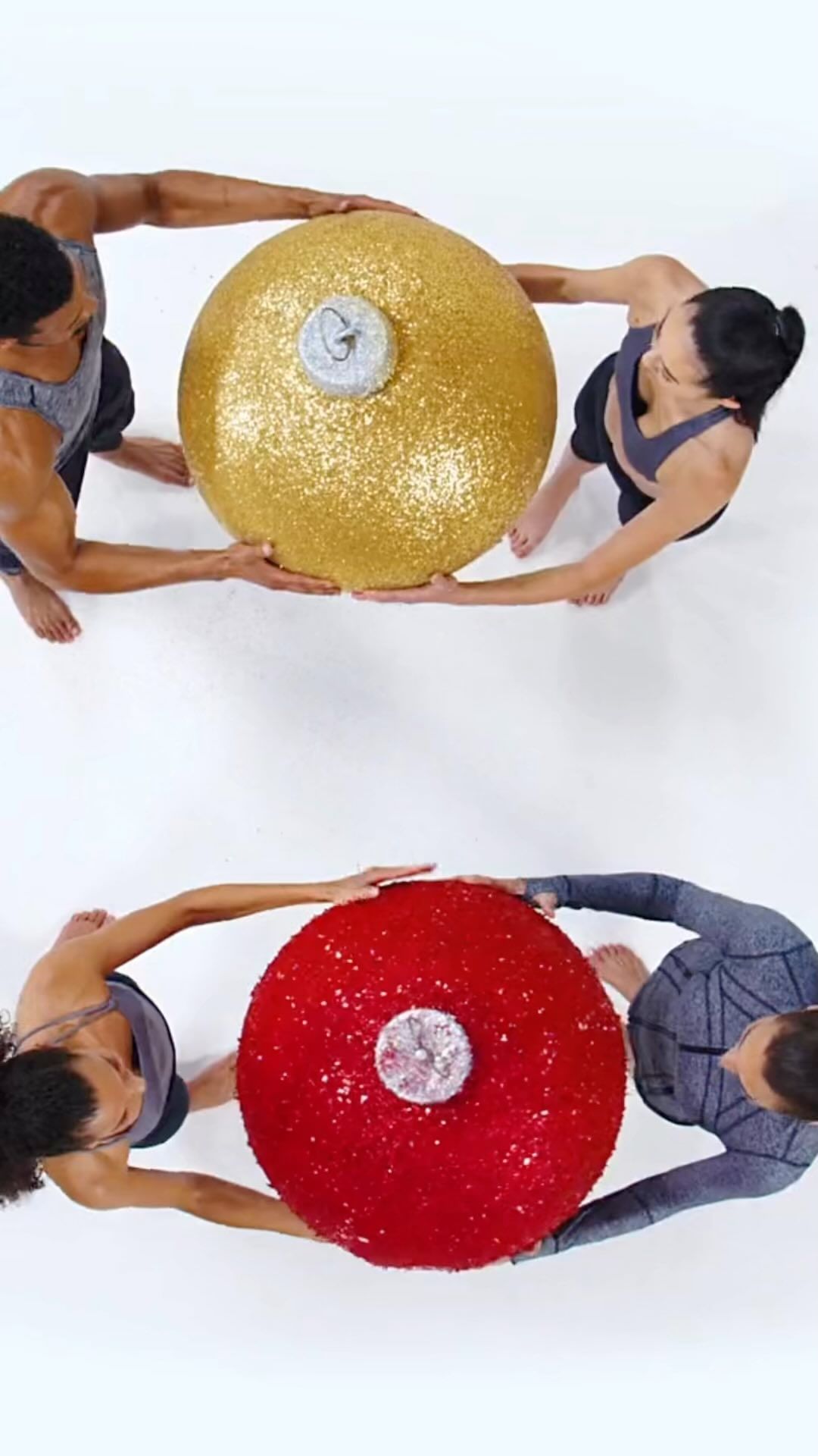 Four athletic adults in fitness wear hold up large, shiny, gold and red Christmas ornaments from an overhead angle on a white background.