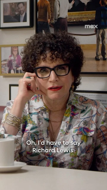 Close-up shot of a woman with curly hair and glasses in a floral shirt sitting at a table with framed pictures on the wall.