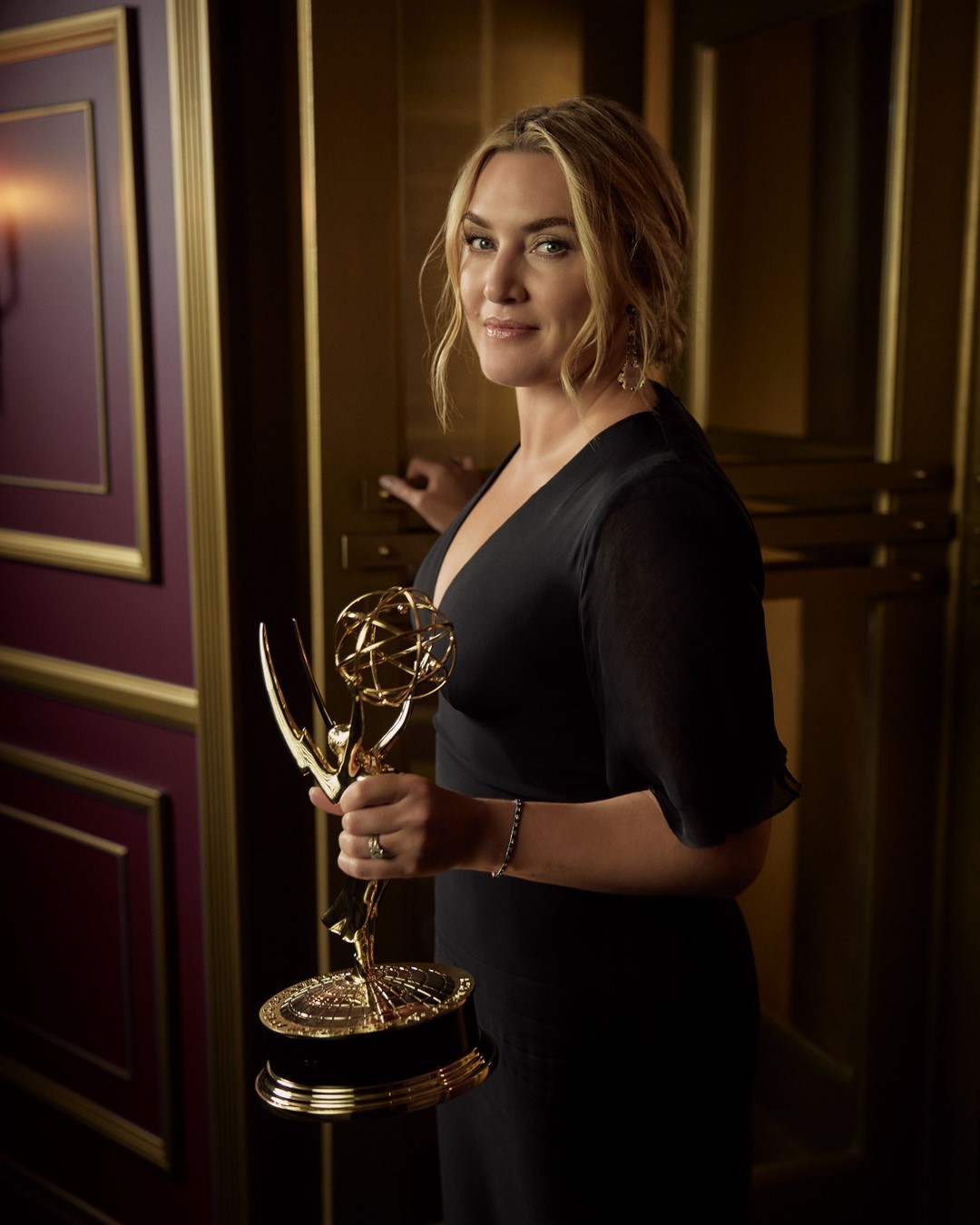 Portrait of Kate Winslet holding an Emmy award in a gold-accented interior setting, wearing a dark dress and soft hair, looking at the camera.