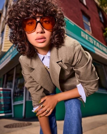 Woman with curly dark hair and amber aviator sunglasses wearing a beige blazer over a striped shirt and blue jeans in New York.