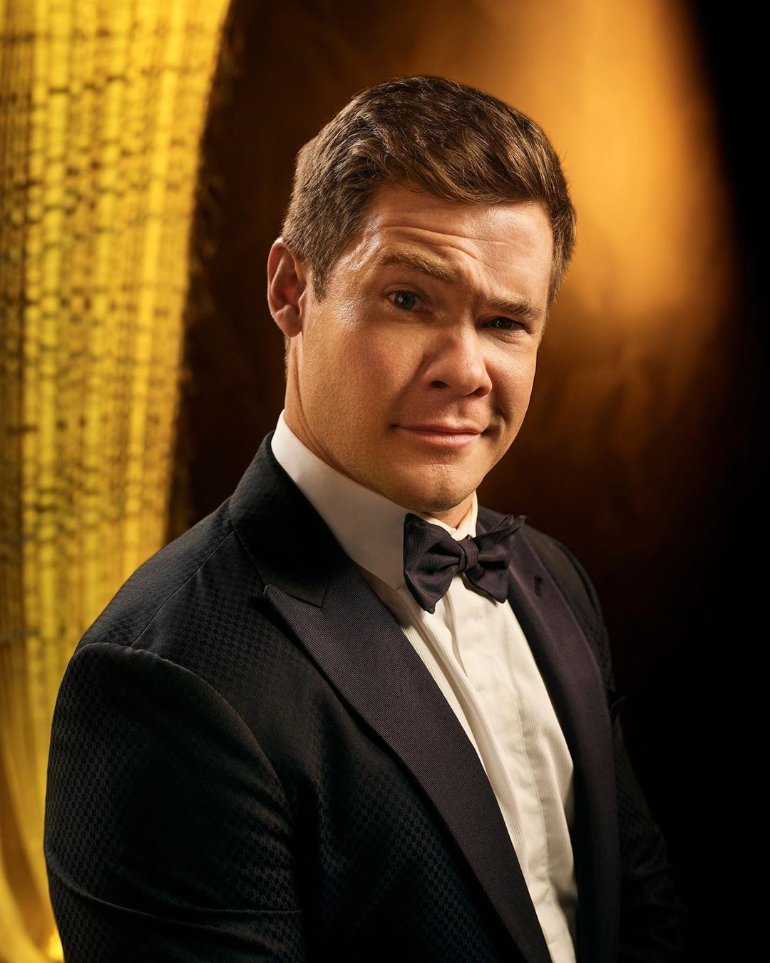 Portrait of adult male in black tuxedo with a bow tie against a gold curtain backdrop, smiling slightly and looking directly at the camera.