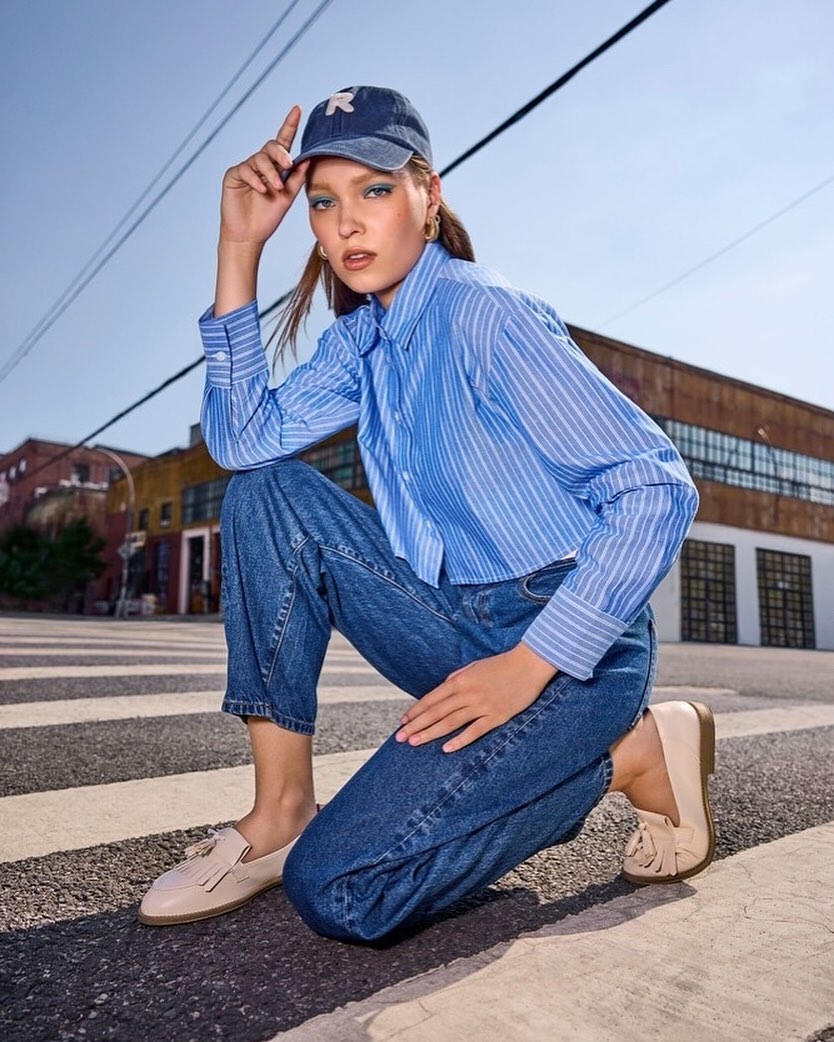 A stylish woman in a blue and white striped shirt and jeans poses on a crosswalk, accessorized with a cap and loafers.