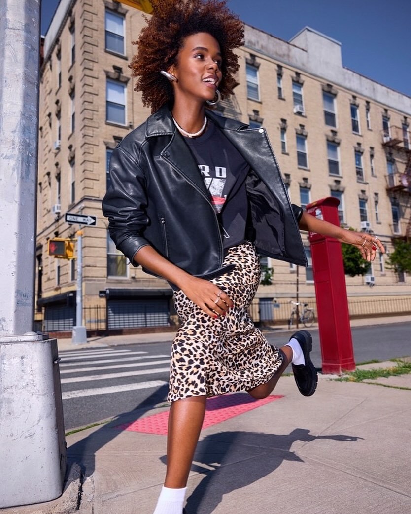 A fashionable African American woman runs on a city sidewalk in a black leather jacket, leopard print skirt, and white socks near a crosswalk.