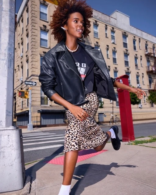 A fashionable African American woman runs on a city sidewalk in a black leather jacket, leopard print skirt, and white socks near a crosswalk.