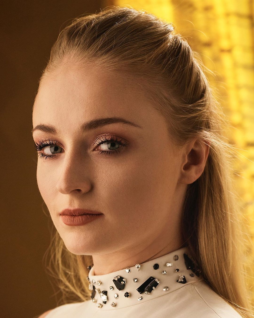 Close-up portrait of an adult woman with blonde hair, wearing a white top adorned with decorative stones, against a golden backdrop.