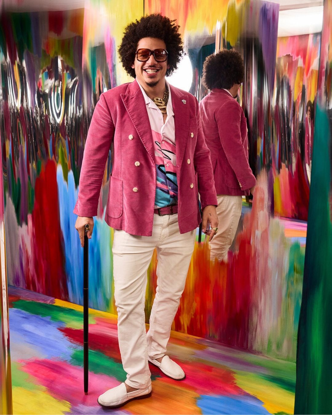 A stylish man with sunglasses, an afro, and a pink blazer poses in front of a colorful wall and reflective panels, holding a cane.