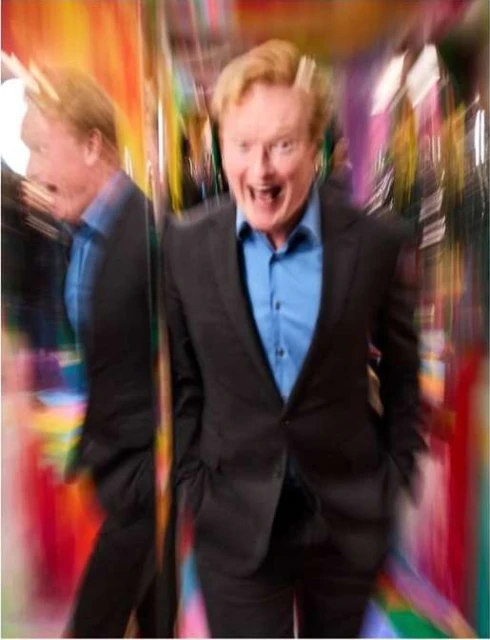 A man in a suit and blue shirt smiles widely with his mouth open, standing in front of a colorful, blurred background with a reflection.