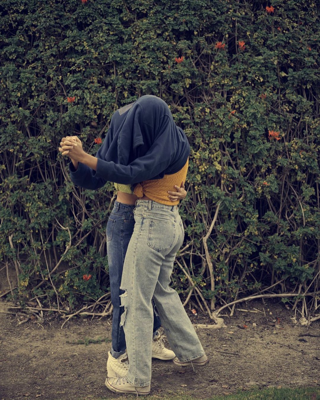 A couple embraces in front of a leafy green hedge with orange flowers, one with a shirt over their head and both in jeans and sneakers.