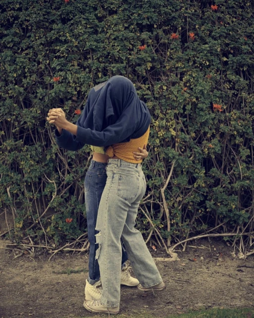 A couple embraces in front of a leafy green hedge with orange flowers, one with a shirt over their head and both in jeans and sneakers.
