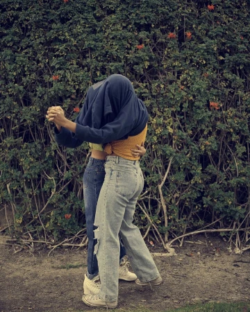 A couple embraces in front of a leafy green hedge with orange flowers, one with a shirt over their head and both in jeans and sneakers.