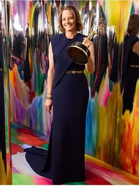 Smiling woman holding an award in a studio with an abstract background, wearing a formal dress with a gold belt and bracelet.