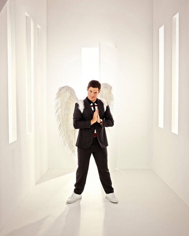 Adult male in suit and angel wings indoors, wearing white shoes with hands clasped. Bright lighting and light walls create a clean space.