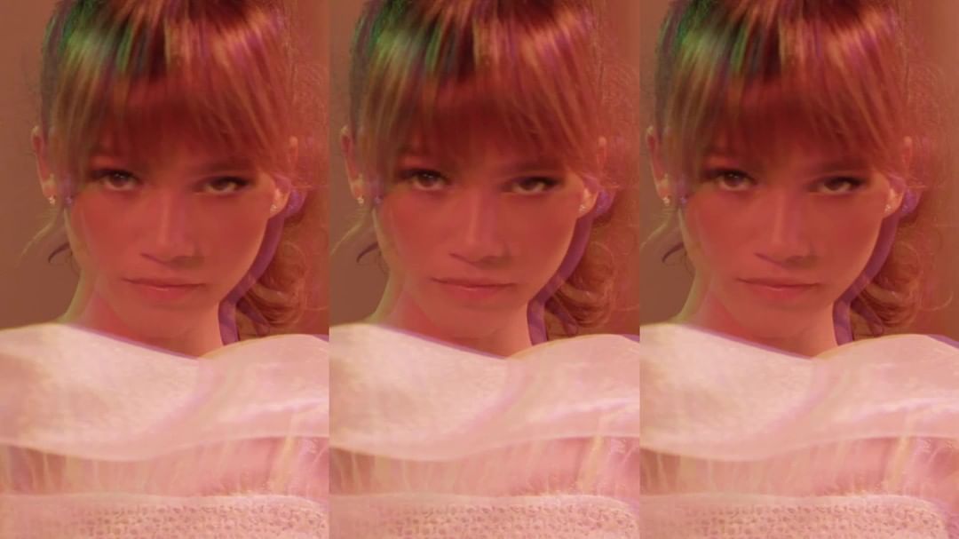 Close-up of a woman with auburn hair, wearing a white frilled top, duplicated across the frame with a pink and green gradient effect.