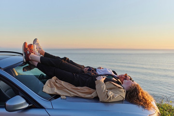 Two people wearing sneakers lie on a silver car with the ocean and horizon in the background at golden hour as the sun sets.
