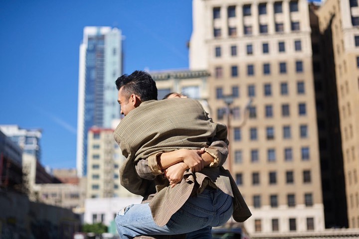Man in blazer hugs someone in urban setting with buildings and blue sky, captured in medium shot, showcasing connection and city backdrop.