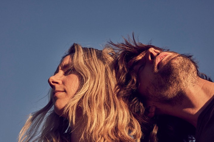 Close-up of a blond woman and brown-haired man leaning back-to-back under a blue sky with soft lighting, portrait orientation.