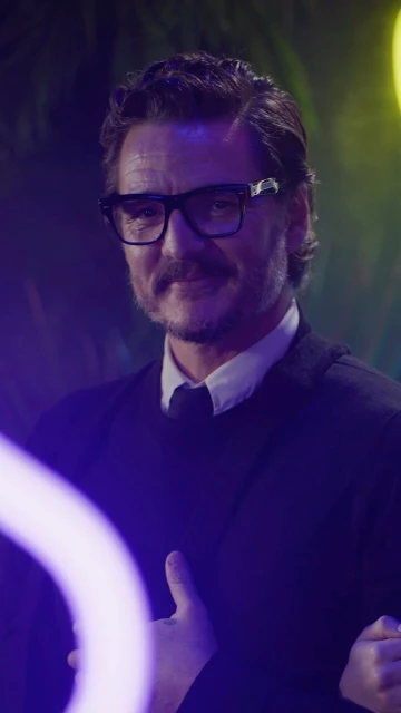 Portrait of actor Pedro Pascal wearing glasses and a dark suit in front of a colorful neon-lit background at an entertainment event.