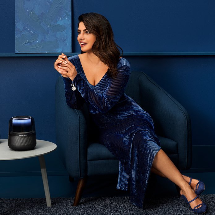 Elegant woman poses in a blue velvet dress and matching heels, seated next to a speaker on a table, set against a blue backdrop.