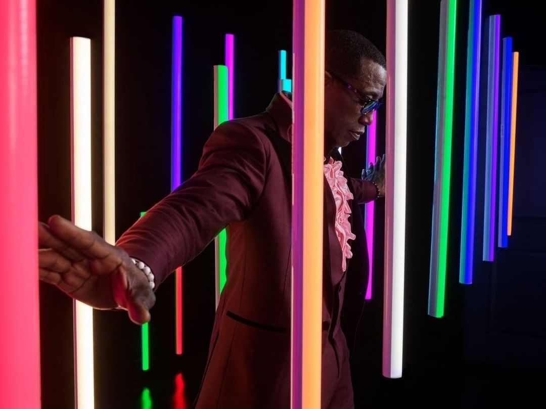 Stylish adult male in glasses and maroon suit stands amidst vertical neon lights, creating a vivid display of color and modern aesthetic.