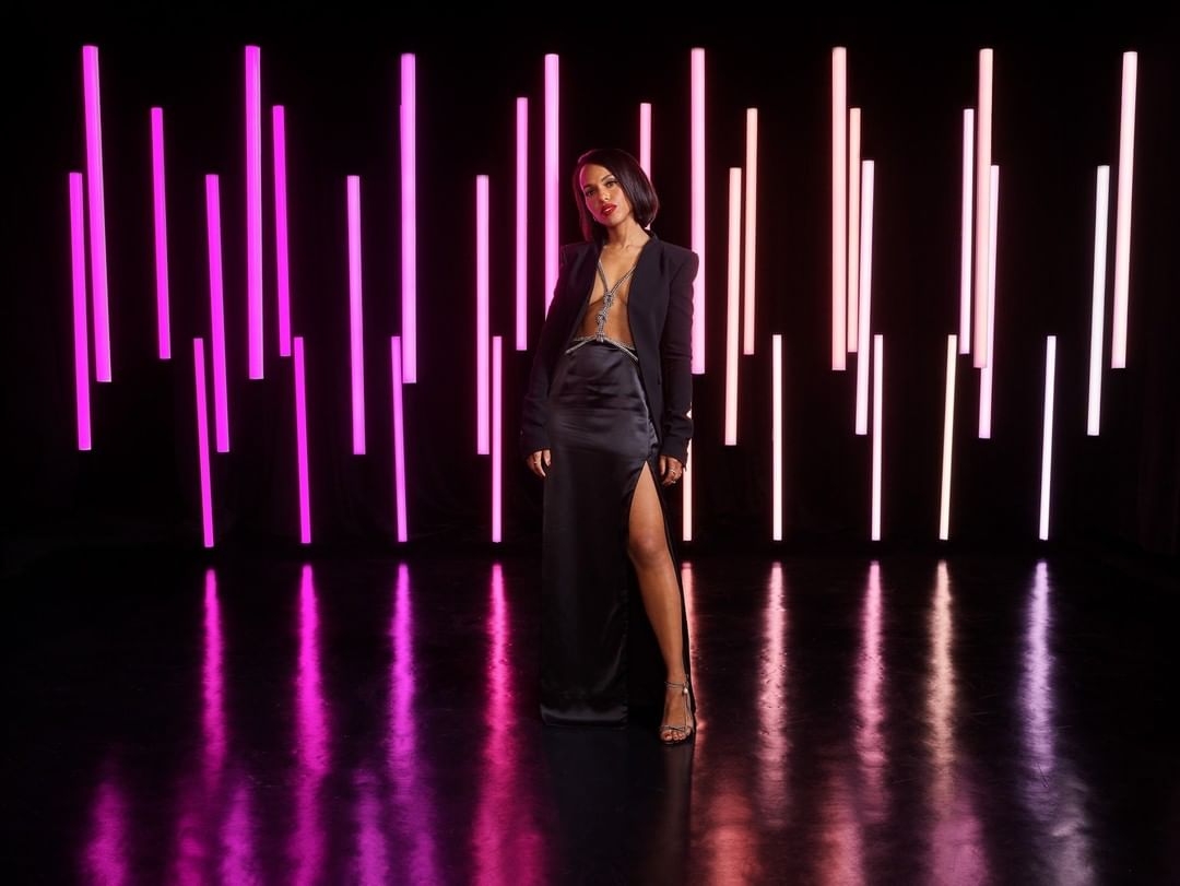 Elegant woman in a black dress and blazer stands in front of vertically arranged neon lights, casting reflections on a black floor.