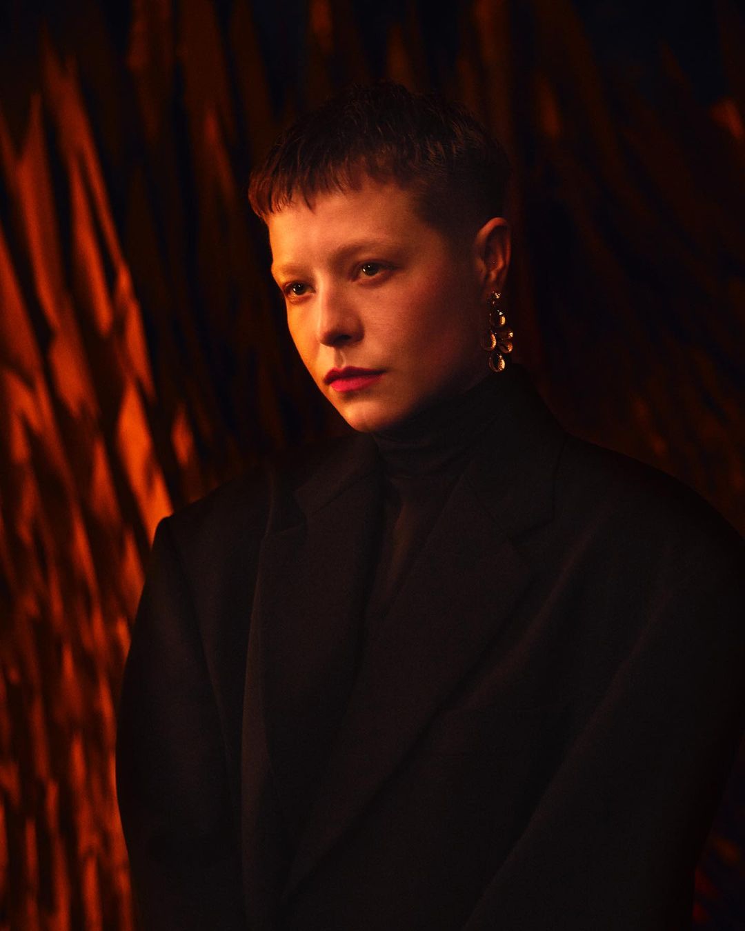 Close-up portrait of a fair-skinned adult female with short hair, wearing a black top and statement earrings against an abstract orange background.