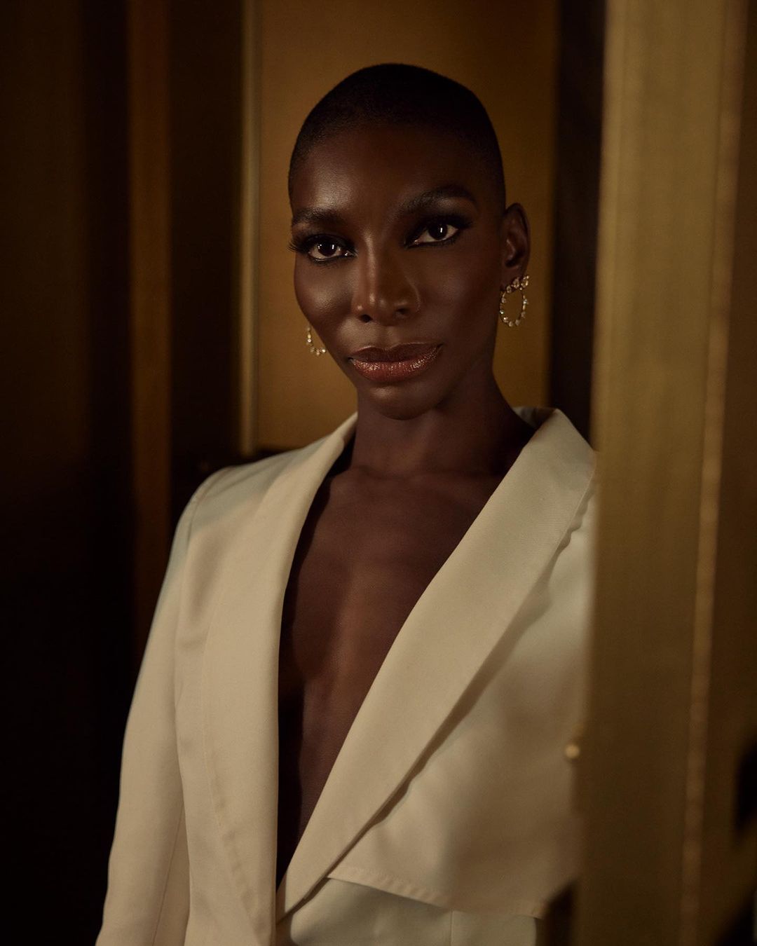 Portrait of a black female in an off-white blazer with short hair, wearing gold hoop earrings against a tan and brown background.