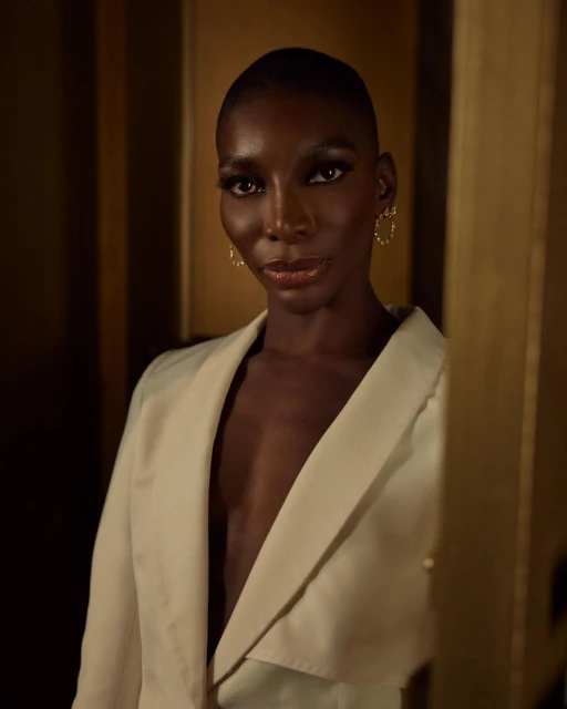 Portrait of a black female in an off-white blazer with short hair, wearing gold hoop earrings against a tan and brown background.