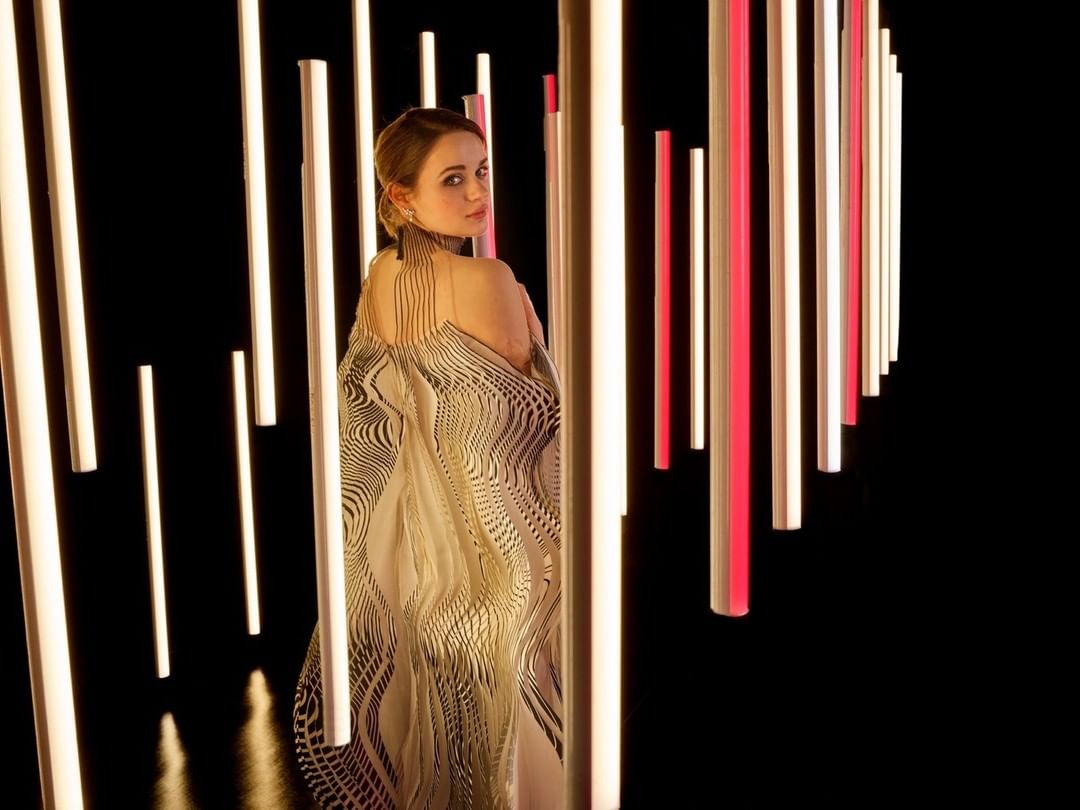 A woman with blonde hair poses in front of vertical fluorescent lights wearing a patterned dress, looking over her shoulder at the camera.