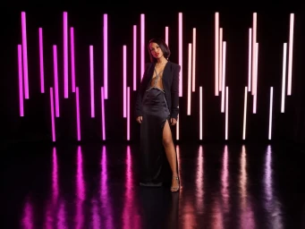 Woman in a black blazer and slit satin skirt posing against a wall of vertical glowing neon pink and white light strips.