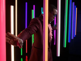 Man in a burgundy suit and pink shirt posing among vertical neon light tubes by Mark Leibowitz