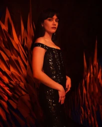 A woman in a black sequin off-the-shoulder dress standing before a dark background with fiery orange spikes.