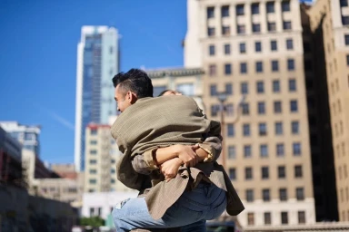 Man holding a woman in his arms on a city street with tall buildings in the background.