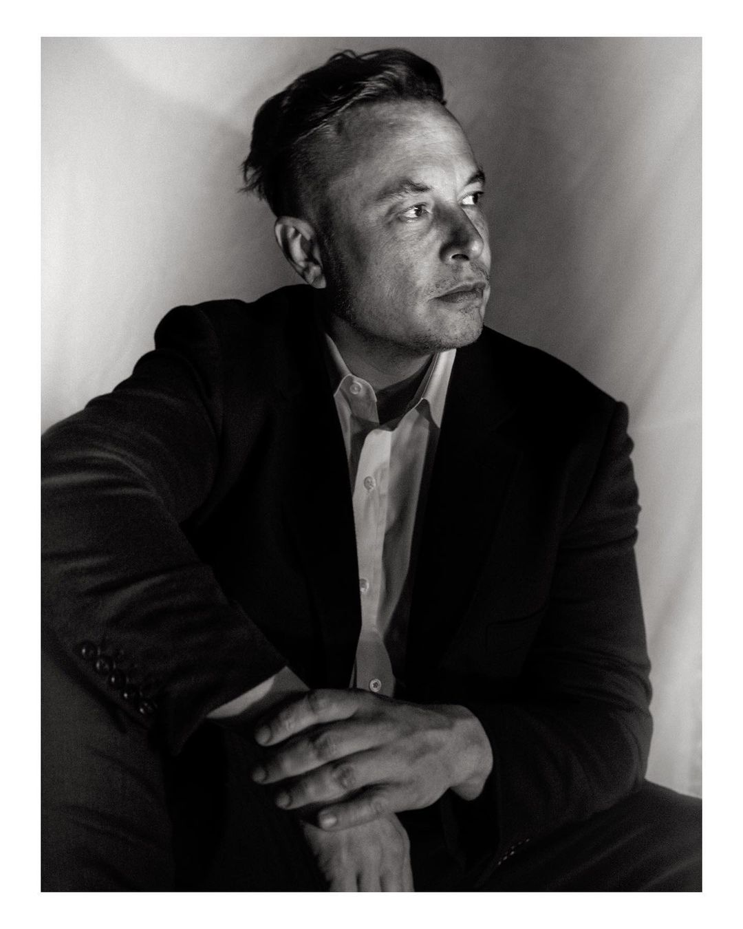 Black and white portrait of Elon Musk in a suit and light shirt, seated with hands clasped, looking off to the side in a photo studio.