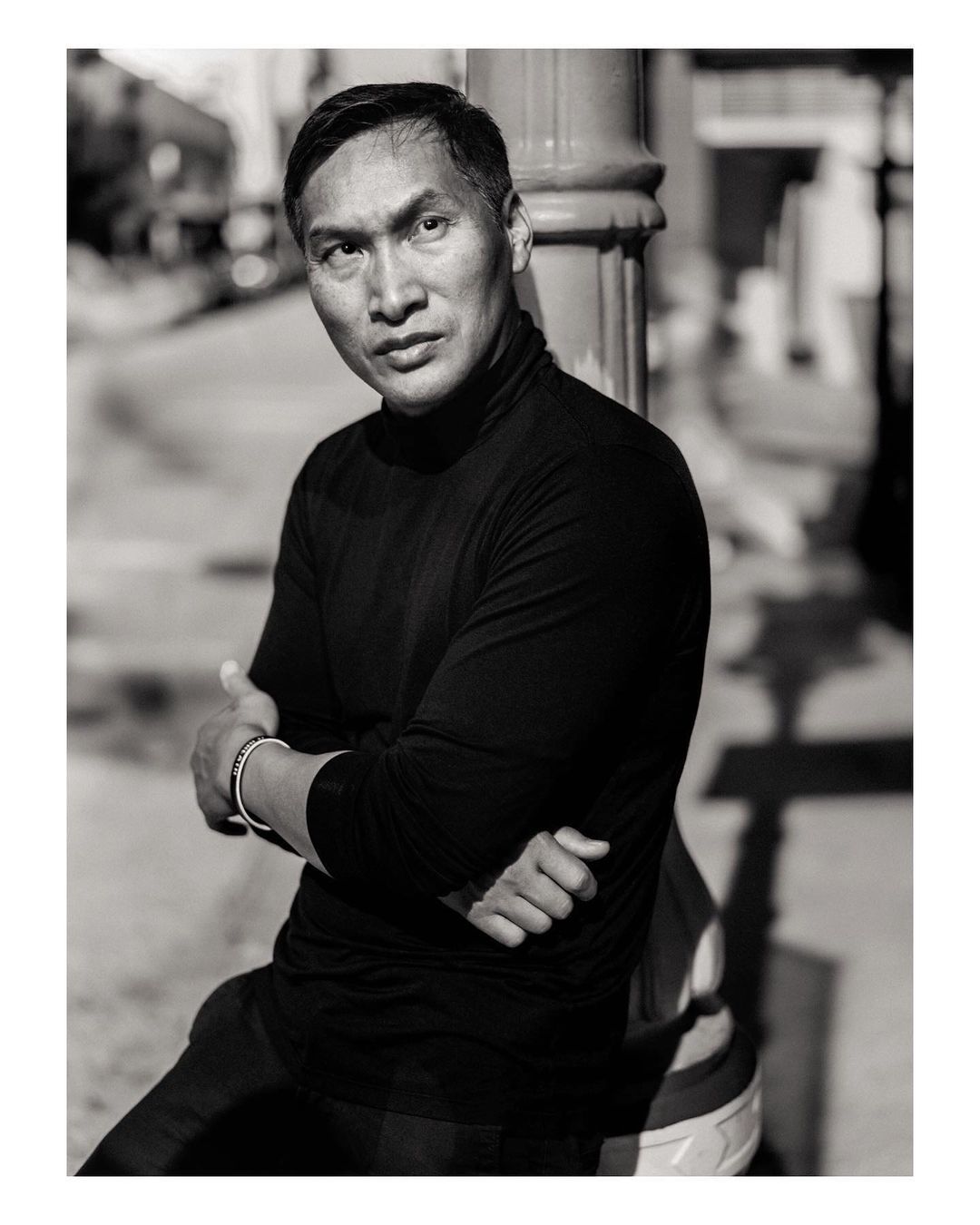 Monochrome portrait of a man with arms crossed, leaning against a lamppost outdoors, showcasing a serious expression and confident posture.