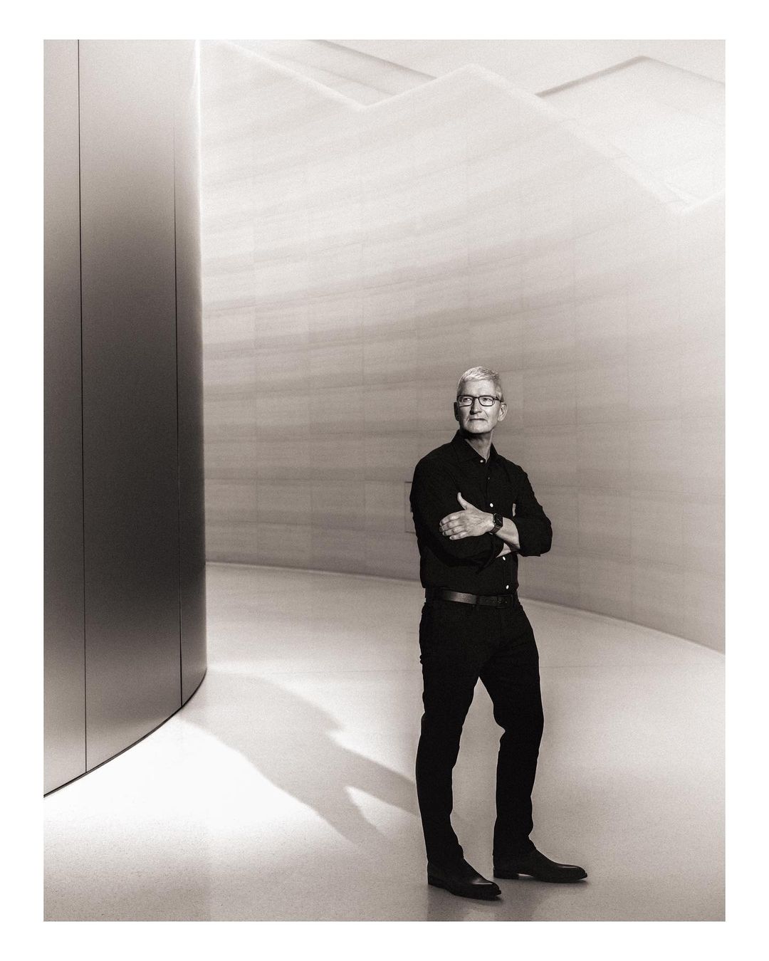 Black and white portrait of a man with arms crossed, wearing a dark shirt and pants, standing in front of an architectural wall.