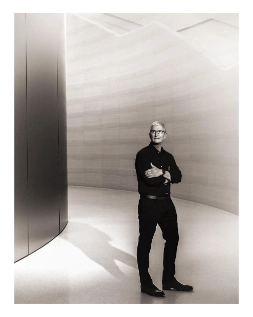 Black and white portrait of a man with arms crossed, wearing a dark shirt and pants, standing in front of an architectural wall.
