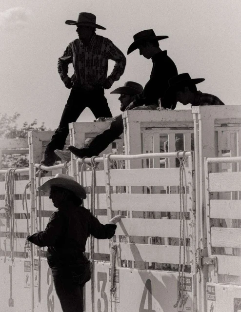 Black and white shot of a group of cowboys in hats and Western wear standing near numbered gates at a rodeo or similar event.