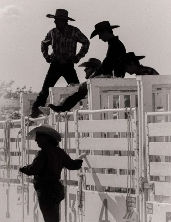 Black and white shot of a group of cowboys in hats and Western wear standing near numbered gates at a rodeo or similar event.